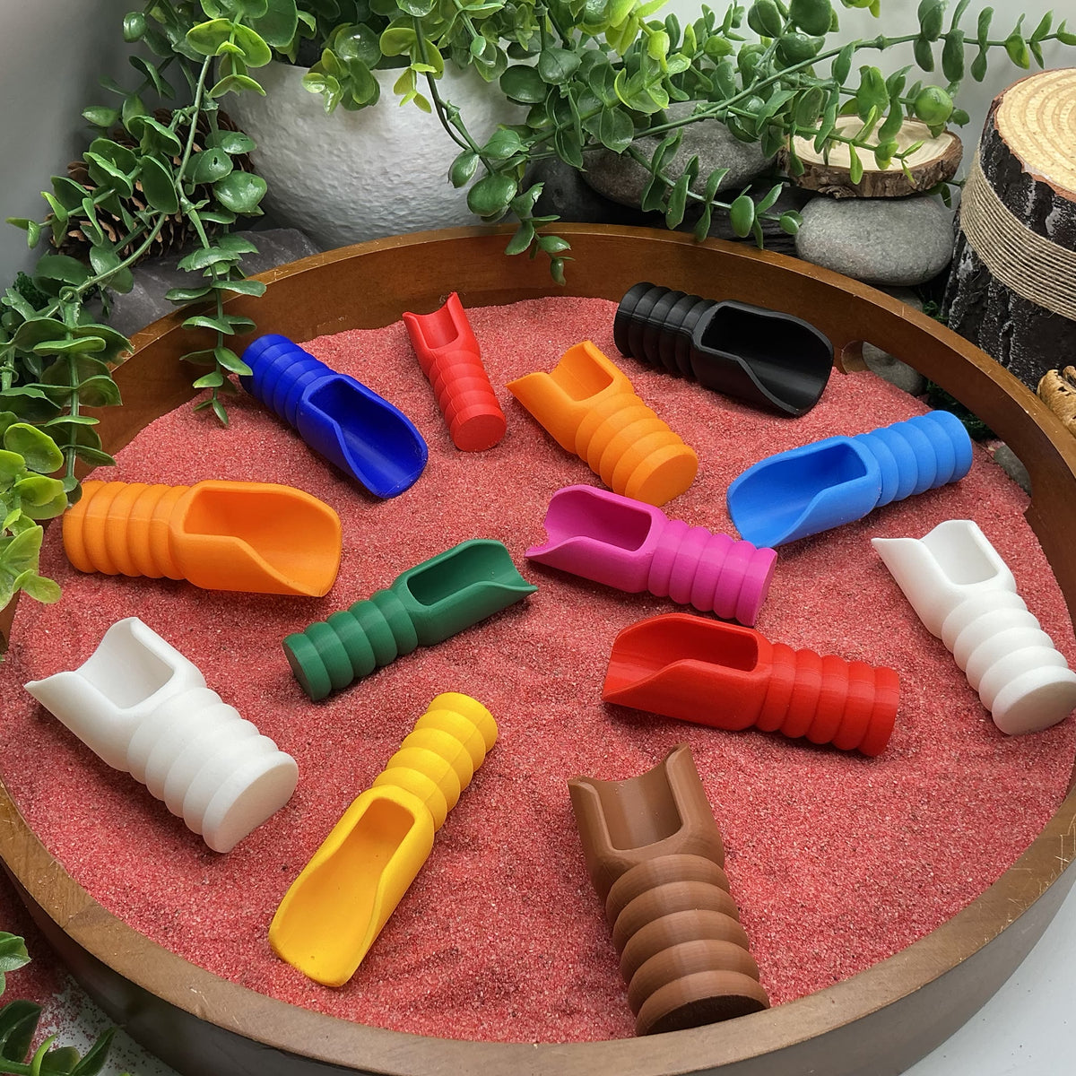 3D printed pouring scoops in a range of colours laid out in red play sand